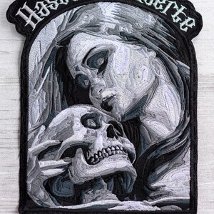 May include: Black and white embroidered patch featuring a skull and a woman's face. The text "Hasta La Muerte" is embroidered around the top of the patch.