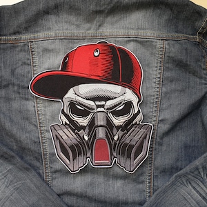 Puede incluir: Una calavera en blanco y negro con una gorra de béisbol roja y una máscara de gas. La calavera está bordada en una chaqueta vaquera.