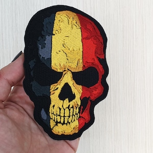May include: Embroidered patch of a skull with the colours of the Belgian flag. The skull is black with yellow teeth and eyes, and the flag is black, yellow and red.