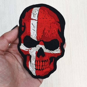 Puede incluir: Parche bordado de una calavera con los colores de la bandera danesa, rojo y blanco, sobre fondo negro.