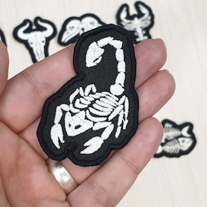 Può includere: Toppa in tessuto nero con uno scheletro di scorpione ricamato in bianco.