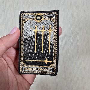 Puede incluir: Parche bordado con la carta del tarot de los Cuatro de Espadas. El diseño incluye cuatro espadas doradas sobre un fondo negro, con un cielo nublado y el título de la carta en texto blanco. El parche tiene un borde dorado.