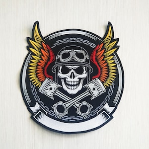 Pode incluir: Um remendo bordado preto e branco com uma caveira usando um capacete e óculos. A caveira é cercada por duas asas com chamas vermelhas, laranja e amarelas. As asas são cruzadas por dois pistões com um design de elo de corrente ao redor do exterior.