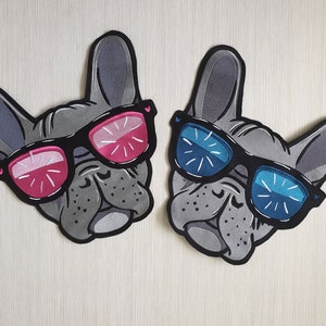 Può includere: Due toppe di feltro grigio di bulldog francesi che indossano occhiali da sole. Un bulldog indossa occhiali da sole rosa e l'altro occhiali da sole blu.