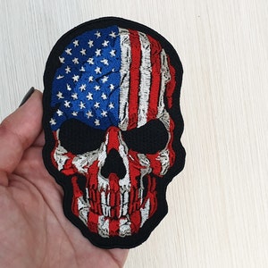 Può includere: Una toppa di teschio ricamata nera con un design di bandiera americana. Il teschio è bianco con dettagli rossi e blu.