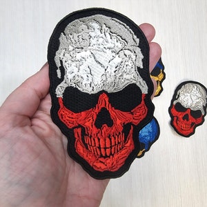 May include: An embroidered skull patch in red, white and black with a skull design. The skull is outlined in black and has a white top with a red and black bottom.