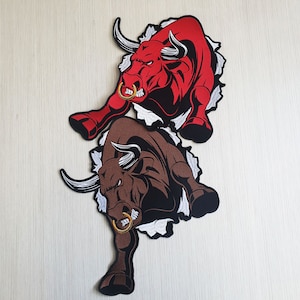 Puede incluir: Dos parches bordados de toros cargando. El toro de arriba es rojo y el toro de abajo es marrón. Ambos toros están rompiendo a través de un fondo blanco.