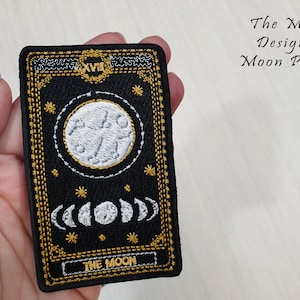 May include: Black embroidered patch with a gold border. The patch features a white moon with a crescent moon and stars. The text "The Moon" is at the bottom of the patch.