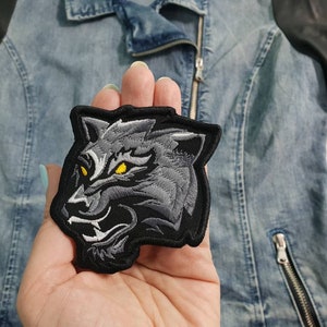May include: Black and grey embroidered patch of a wolf's head with yellow eyes.