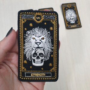 May include: Black and gold embroidered tarot card with a white lion and skull design. The card reads "VIII STRENGTH".