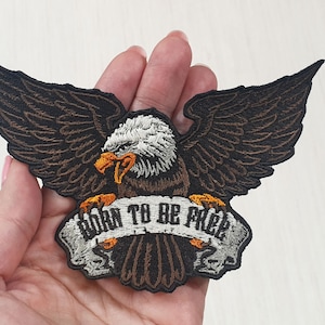 Peut inclure: Patch brodé représentant un aigle brun avec une tête blanche et un bec jaune, tenant une bannière avec le texte "Born to be Free".