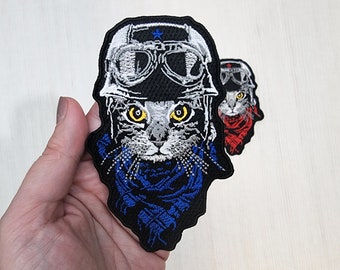 Puede incluir: Parche bordado para planchar con un gato que lleva un casco negro con gafas y una bufanda azul. El gato tiene ojos amarillos y una nariz blanca.