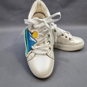 May include: A pair of white leather sneakers with white laces. One shoe has a blue, green, and yellow mountain patch on the side.