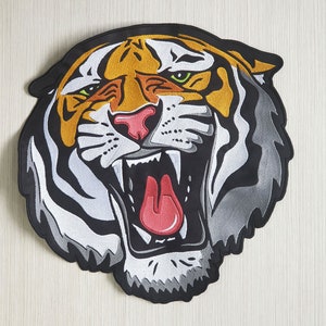 Puede incluir: Un parche bordado de cabeza de tigre negro, blanco y naranja con una expresión de gruñido y una lengua roja.