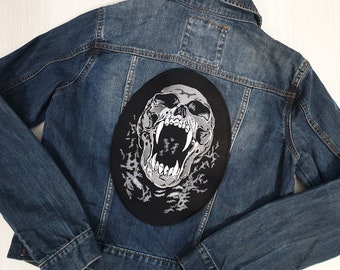 Op de afbeelding: Een zwart denim jack met een wit geborduurde schedel patch op de rug. De schedel gromt met zijn mond open en zichtbare tanden.