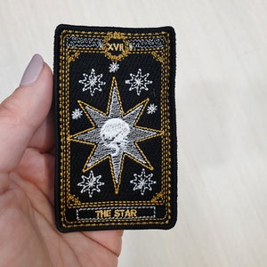 Könnte beinhalten: Schwarzer gestickter Patch mit einem weißen Stern und dem Text "THE STAR" in einem goldenen Rahmen. Der Stern hat einen weißen Kreis in der Mitte mit einem weißen Schädel darin.