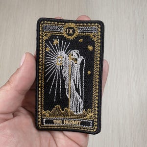 May include: A rectangular embroidered patch featuring a black background with gold and white thread. The design depicts a figure holding a lantern with radiating light, the Roman numeral IX, and the text THE HERMIT in a decorative gold border.