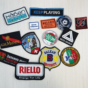 Op de afbeelding: Een verzameling geborduurde patches met verschillende logo's en ontwerpen. De patches zijn kleurrijk en bevatten tekst zoals "Keep Playing", "Bulleit Frontier Whiskey", "GH.MUMM", "Balkan Security" en "Riello Energy For Life".