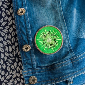 Könnte beinhalten: Eine blaue Jeansjacke mit einem grünen gestickten Kiwi-Frucht-Patch auf der Vorderseite. Die Jacke hat einen Knopfverschluss und ein weißes Blumenmuster auf der Innenseite.