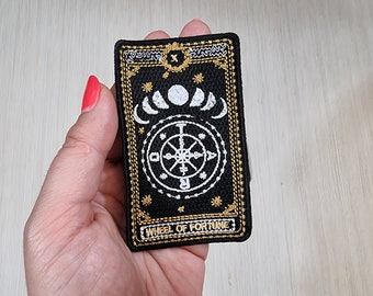 May include: A black and gold embroidered patch with a white wheel of fortune design. The patch features a circle with a cross in the center and four arrows pointing outward. The text "WHEEL OF FORTUNE" is printed below the wheel.