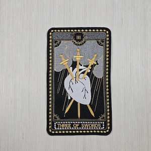 May include: Embroidered patch featuring the Three of Swords tarot card. The design includes a white heart pierced by three gold swords, set against a stormy gray sky. The card is framed in black with gold accents, and the text "THREE OF SWORDS" is at the bottom.