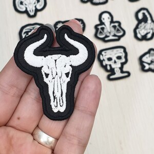 Könnte beinhalten: Weiß gestickter Stier Schädel auf einem schwarzen Stoff-Patch. Der Patch wird in einer Hand gehalten.
