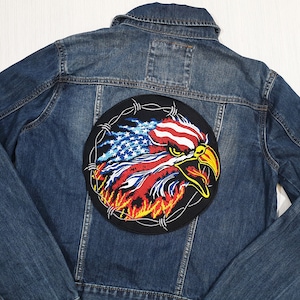 Può includere: Una giacca di jeans con una toppa rotonda ricamata che raffigura un'aquila con un design di bandiera americana. L'aquila è circondata da filo spinato.