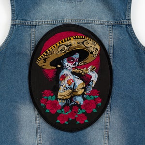 Op de afbeelding: Een geborduurde patch in zwart en rood met een vrouw met een schedelgezicht die een grote sombrero draagt en een bloem vasthoudt. De vrouw wordt omringd door rode rozen. De patch zit op een blauwe denimvest.