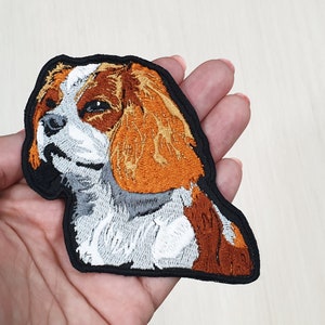 Puede incluir: Parche bordado para planchar de un perro Cavalier King Charles Spaniel con pelaje marrón, blanco y naranja. El parche está sobre un fondo negro.