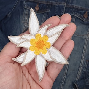 May include: A white embroidered flower patch with a yellow centre. The flower has five petals and is a popular symbol of the Alps.