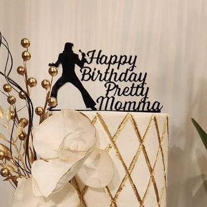 May include: A white cake with gold diamond pattern and a black silhouette cake topper that reads "Happy Birthday Pretty Momma." The cake is decorated with white flower accents and gold decorative branches.