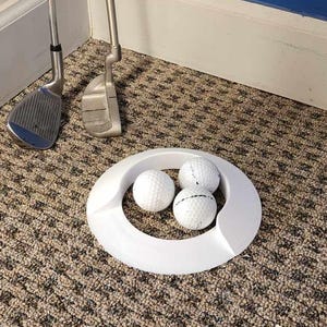 May include: A white golf ball training aid with three white golf balls inside. The aid is on a brown carpet.