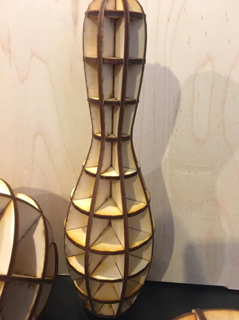 Pode incluir: Um vaso de madeira em forma de pino de boliche com um design geom&eacute;trico. O vaso &eacute; feito de v&aacute;rias pe&ccedil;as de madeira que s&atilde;o unidas para criar uma estrutura de treli&ccedil;a. O vaso &eacute; de cor marrom claro e tem um acabamento de madeira natural.