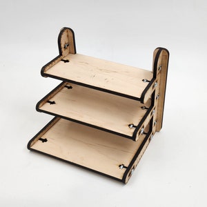 Op de afbeelding: Een houten plank met drie niveaus, met een natuurlijke afwerking en zwarte randen. De planken worden bij elkaar gehouden met metalen hardware. Het ontwerp is eenvoudig en functioneel, geschikt voor het uitstallen van kleine items of het organiseren van benodigdheden.