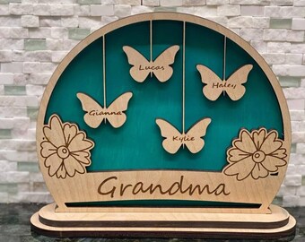 Op de afbeelding: Houten uitsnijding van een teal cirkel met vier vlinders die eraan hangen. Elke vlinder heeft een naam: Lucas, Haley, Gianna en Kylie. De cirkel is omgeven door twee bloemen en het woord "Oma" staat onder de cirkel.