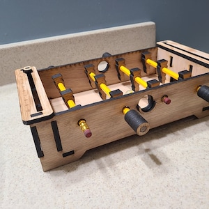 Puede incluir: Un juego de futbolín de mesa de madera con lápices amarillos como jugadores y pomos negros para las asas. El juego está hecho de madera y tiene un acabado natural.