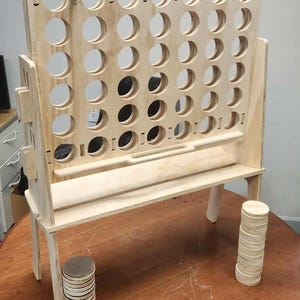 May include: A wooden Connect Four game set. The game board has a grid of circular holes, supported by a stand. Two stacks of wooden game pieces are on the table. The game is made of light-colored wood.