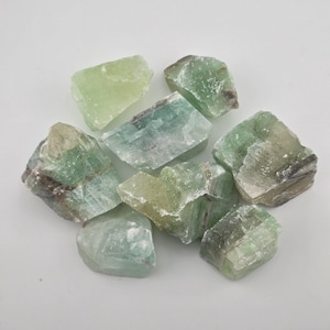 May include: A collection of raw, natural green fluorite crystals. The crystals vary in shades of green, from pale to deep, with some showing hints of blue and white. The rough, irregular shapes of the stones are visible.