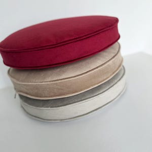 May include: A stack of three round, flat cushions with a soft, velvet-like texture. The top cushion is deep red, the middle cushion is a light beige, and the bottom cushion is a light gray. Each cushion features a visible side zipper.