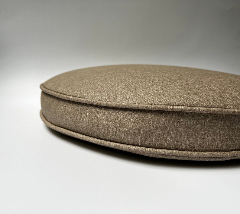 May include: A round, light brown cushion with a textured fabric and a stitched edge.