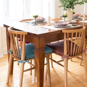 May include: A wooden dining table with four spindle-back chairs featuring tie-on seat cushions in blue and mauve. The table is set with white placemats, grey bowls, green wine glasses, white candles, and a vase with green foliage.