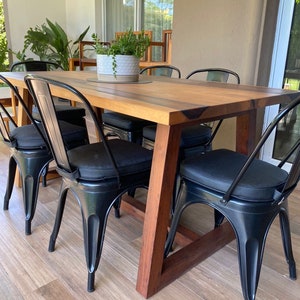 Puede incluir: Una mesa de comedor de madera con un acabado de madera natural y sillas de metal negro. La mesa tiene forma rectangular y está rodeada por seis sillas. Las sillas tienen marcos de metal negro y cojines negros.