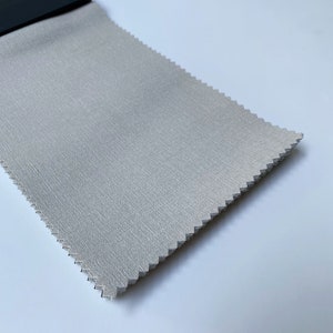 May include: A close-up of a light gray fabric swatch with a subtle textured pattern. The fabric is folded and appears to be a sample for upholstery or drapery.