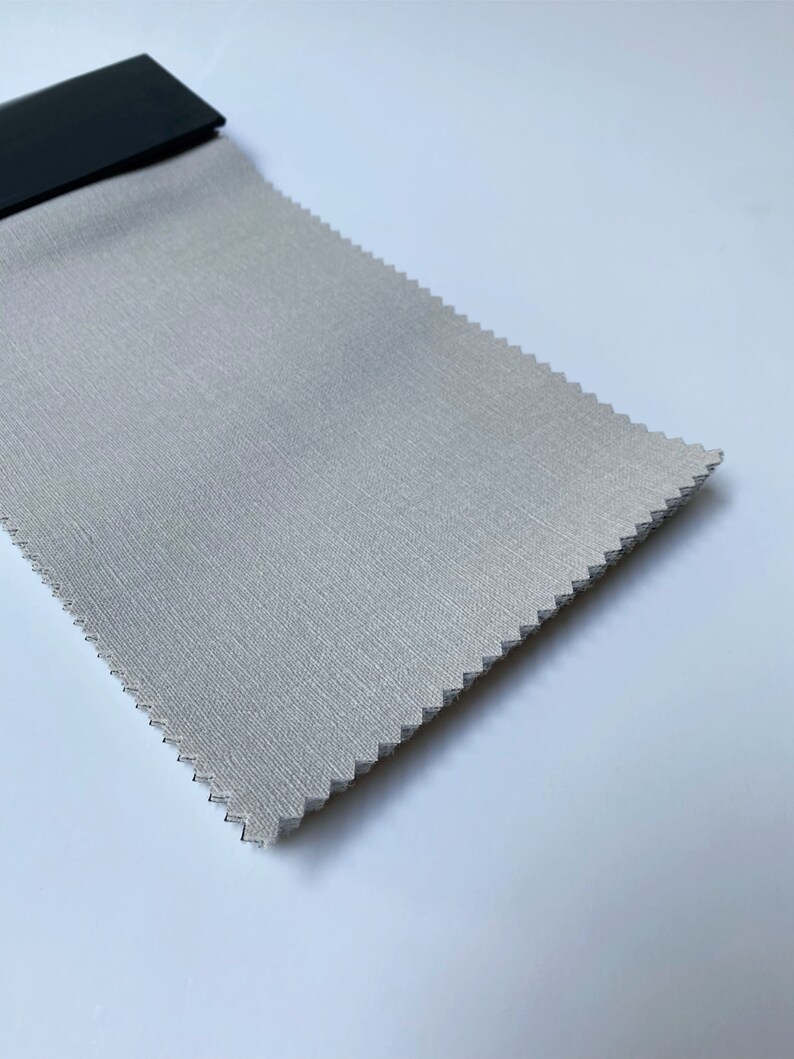 May include: A close-up of a light gray fabric swatch with a subtle textured pattern. The fabric is folded and appears to be a sample for upholstery or drapery.