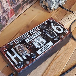 Op de afbeelding: Een handgemaakte gitaar van een sigarenkistje met een Route 66 nummerplaat ontwerp. De gitaar heeft een houten hals en een zwart-zilveren gevlochten kabel. Een luidspreker met een slangenprint en een bord met "GOOD FOOD COLD BEER LOUD MUSIC" staan op de achtergrond.