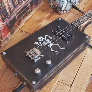 Op de afbeelding: Een handgemaakte sigarenkistgitaar met een zwarte body en een skeletafbeelding. De gitaar heeft een lange hals, snaren en een zilveren brug. Een luidspreker met slangenpatroon en een vintage bord staan op de achtergrond. De tekst "CigarBoxMusic.fr" staat rechtsonder.