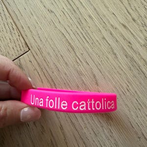 Può includere: Un braccialetto in silicone rosa acceso con la scritta bianca "Una folle cattolica". Il braccialetto è mostrato su uno sfondo di legno marrone chiaro.