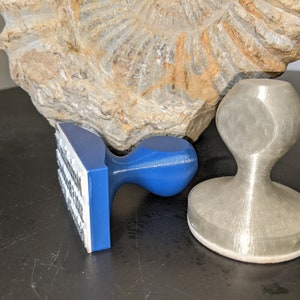 May include: Two different rubber stamps, one blue and one silver. The blue stamp has a rectangular base and a rounded handle. The silver stamp has a round base and a rounded handle. Both stamps are on a dark surface.