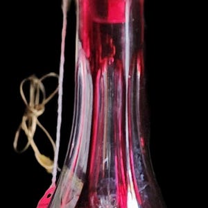 May include: A ruby red glass decanter with a clear, faceted stopper. The decanter features intricate cut glass designs, including starburst and floral patterns. A tag is attached to the neck. The background is black.