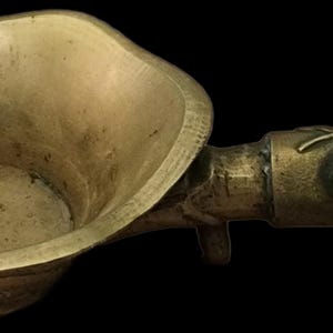 May include: A vintage brass oil lamp with a shallow bowl and a long handle. The handle is decorated with a leaf design, a red stone, and a green stone. The lamp has an aged, antique appearance.